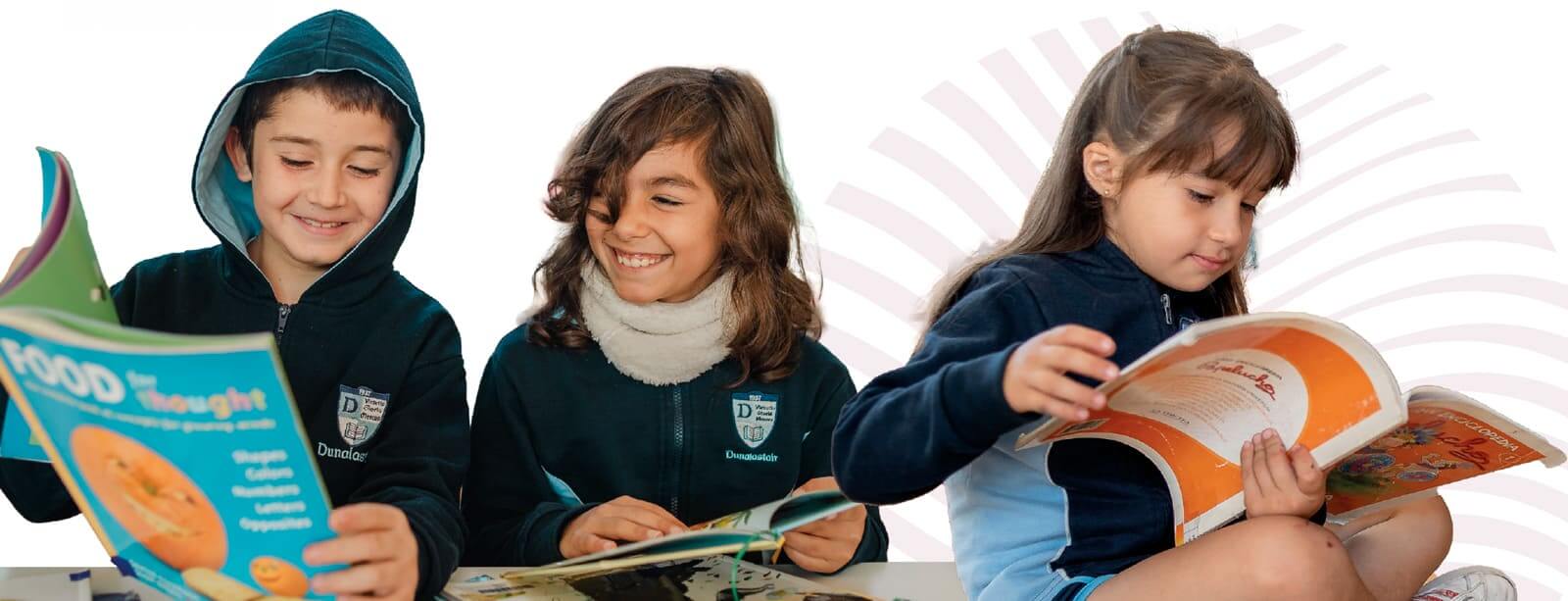Tres niños sonrientes en edad escolar, vestidos con uniformes azul marino y azul claro que llevan el logotipo de Dunalastair School, sentados y leyendo libros de cuentos coloridos. Un niño con una sudadera con capucha abierta sostiene un libro titulado "FOOD for thought", otra niña a su lado sonríe mientras lee, y una tercera niña a la derecha lee su propio libro, todo sobre un fondo blanco limpio con un sutil patrón geométrico rosa. Imagen que muestra la alegría de la lectura infantil y el entorno de aprendizaje en Dunalastair School.